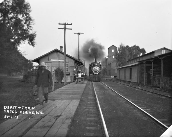 View down railroad tracks of Cross Plains train depot. A man in the foreground walks away from the depot. A train, exhausting steam, is stopped at the platform. Lumber is stored in a building across from the depot.