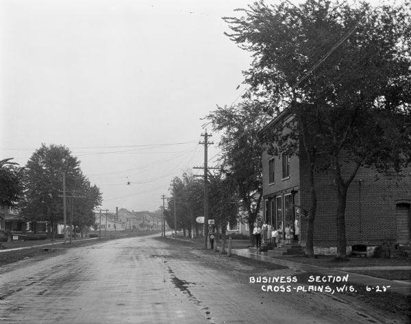 View down road of mixed residential and commercial area after a rain. A group of men, women, and children stand on the front steps of a brick building.