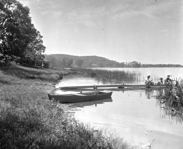 View along shoreline of a man and two boys fishing on a wooden dock. A rowboat is pulled up on the shore. Two cars are parked near trees. In the far background are hills and farm buildings.