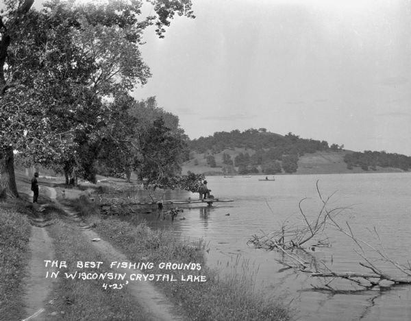 View down dirt road along the shoreline of Crystal Lake. A man and a child stand under a tree's shade, near a pier where a man is seated on the dock. There are boaters in the lake beyond the pier. In the far background is a hill.