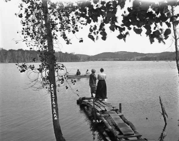 View from shoreline of two women standing on a dock on Crystal Lake. Just beyond the dock, a man and woman fish from a boat.