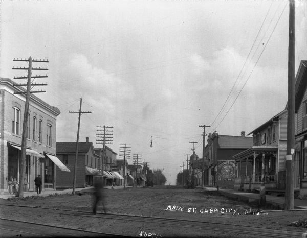 View down commercial Main Street, looking north. A pedestrian (blurred from movement) is crossing the street along what appear to be railroad tracks. There is a painted advertisement for Richards and Buffington beer by the Dubuque  Brewing and Malting Company. Pedestrians are on the sidewalk, and a man is driving a horse-drawn wagon.