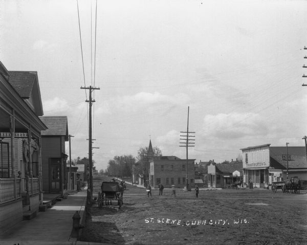View along sidewalk of scene of street where children and an adult stand near railroad tracks. Donohoo Splinter & Company is across the street, as well as a Pool Room. Four horse-drawn vehicles are parked or traveling along the street.