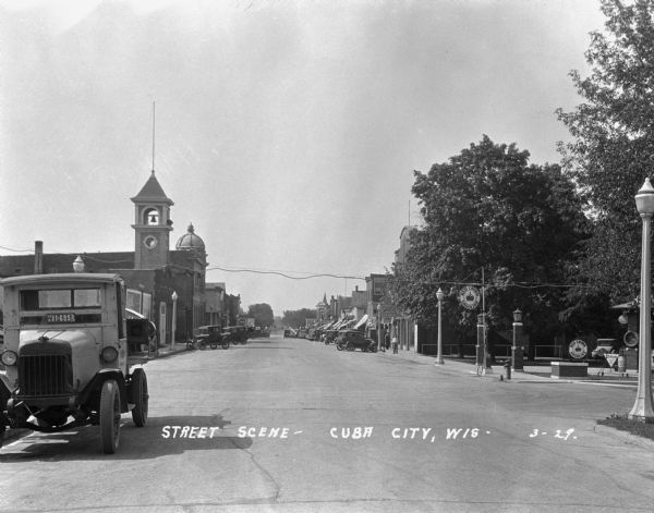View down a commercial street. An International truck is parked in the foreground on the left, and on the corner on the right is the Red Crown Gasoline station. In the background cars are parked at an angle in front of storefronts.