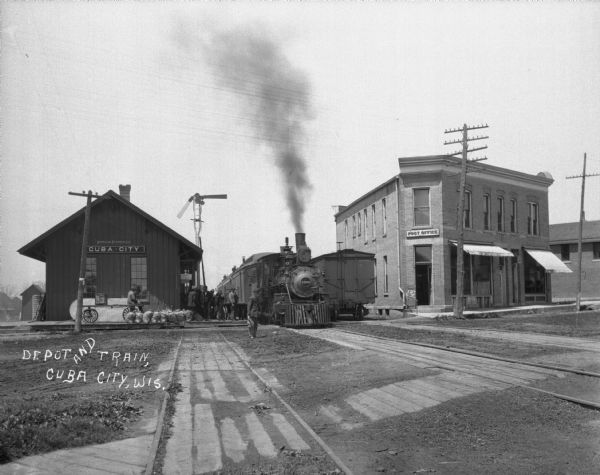 View down railroad tracks of a busy scene at the train depot. A train is arriving at the crowded platform. On the side of the depot a man sits on a cart, near sacks propped along the platform. Across from the depot is a post office.