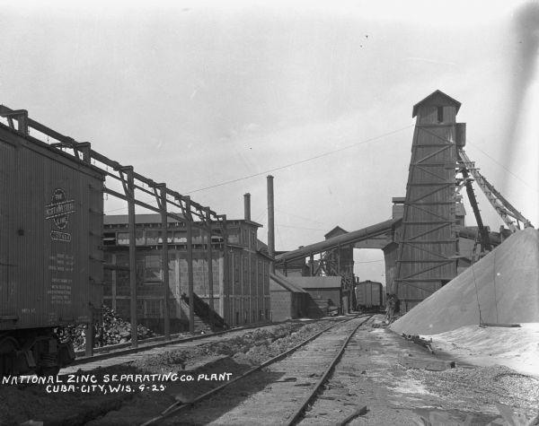 View down railroad tracks of the National Zinc Separating Company grounds. There is a large mound of ores, railroad cars, industrial buildings and a tall building with a chute.