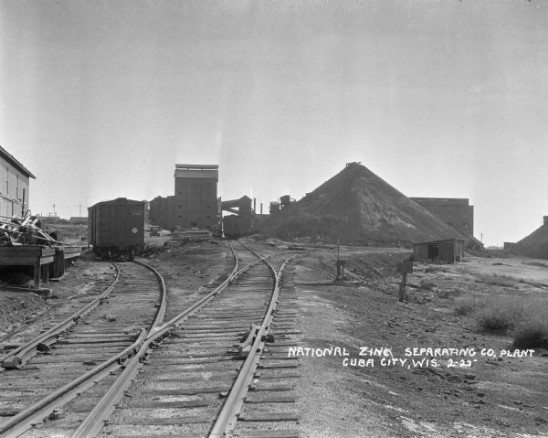 View down railroad tracks towards large mounds of raw material at National Zinc Separating Company plant. Industrial buildings and a railroad car and an automobile are on the left.