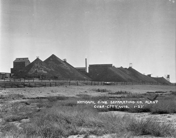 View across field towards large mounds of raw material at National Zinc Separating Company plant. Industrial buildings are in the background.