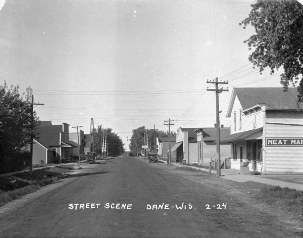 View down a commercial street. There is a meat market on the right, and railroad crossing gates further down the street near other commercial buildings.