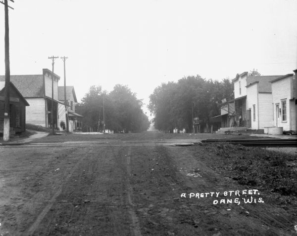 View down middle of street towards the railroad crossing at Military Road. There are shops on the left and right, and trees line the street in the background.