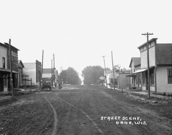 View down the unpaved Military Road. Two boys stand in the street on the left near the State Bank of Dane building. Near the railroad crossing, there is a sign alerting pedestrians to "look out for the cars."