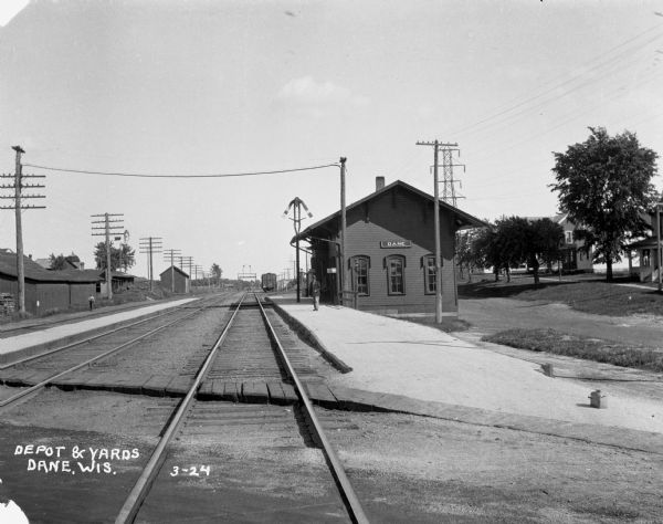 View down railroad tracks of a man standing on the platform at the Dane train depot. Houses are along a street on the right.