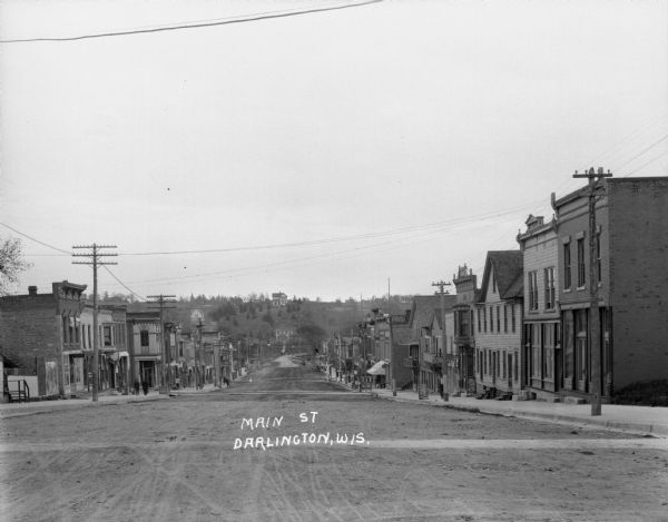 View down Main Street looking south. At the bottom of the street is a bridge, and beyond is a hill with houses and trees. There are pedestrians along the sidewalk of Main Street, which is lined with storefronts.