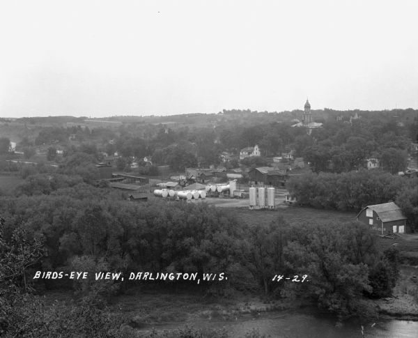 Elevated view of town looking down a hill. In the foreground is a river with trees along the bank. On the other side of the river is a factory with industrial buildings and a number of storage tanks. The Lafayette County Courthouse and houses are in the background among more trees.