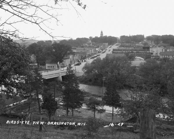 View down tree-lined hill over the Pecatonica River. The bridge connects River Street to Main Street. Storefronts line the street, and the Lafayette County Courthouse and the Soldiers and Sailors of the Civil War monument are in the background.