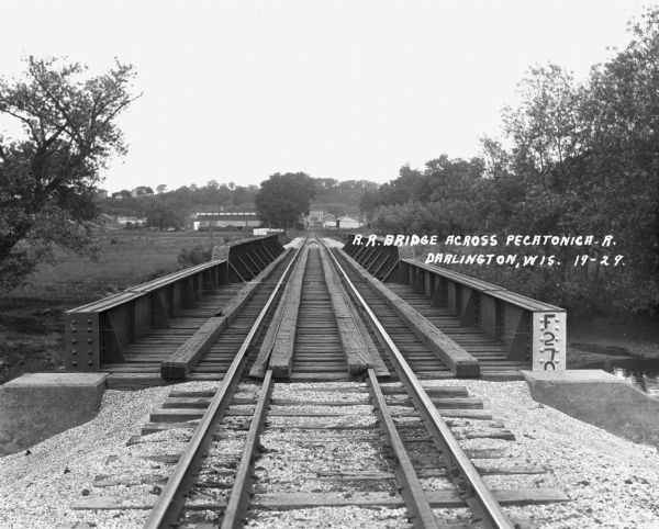 View down the railroad tracks across the bridge. On the far side are barns and industrial buildings among trees.