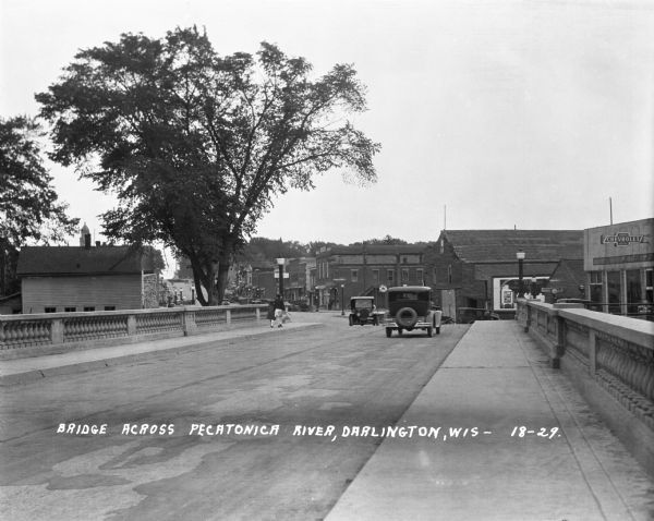 View across the bridge on Main Street. There are two cars and a woman and child walking across the bridge. A Chevrolet dealership is on the right.
