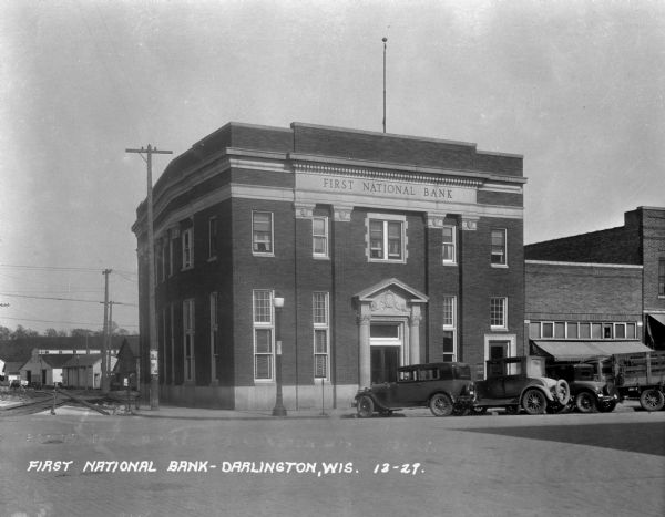 View from across street of the exterior of the First National Bank at 245 Main Street. Automobiles are parked at an angle along the curb in front. On the left are railroad tracks, and in the background are industrial buildings and a ridge.