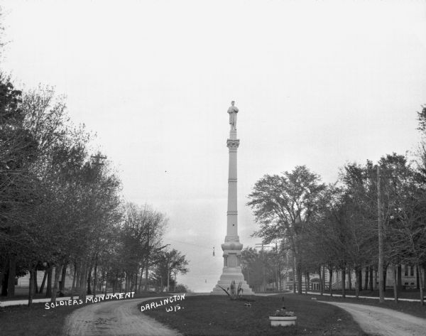 The Soldiers and Sailors of the Civil War memorial on the unpaved Main Street. The memorial features a plaque, a shield, crossed swords, and monument of a Union soldier at parade rest. On the right, there is the high school and the grade school.