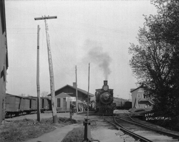 View down railroad tracks of a train at the Darlington depot. A group of men are on the platform.