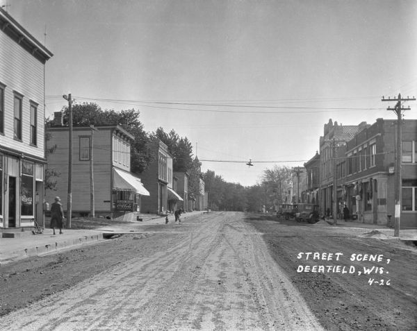 View down an unpaved street at the intersection of Main Street and Nelson Street. There are pedestrians on the sidewalks, and a man is shoveling in the street outside the Nelson Brothers store.