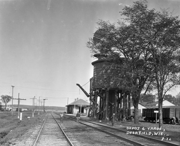 View down railroad tracks where three boys are standing. The water tank and windmill are on the right near the Deerfield Depot. On the far right, two men are pushing a train car.