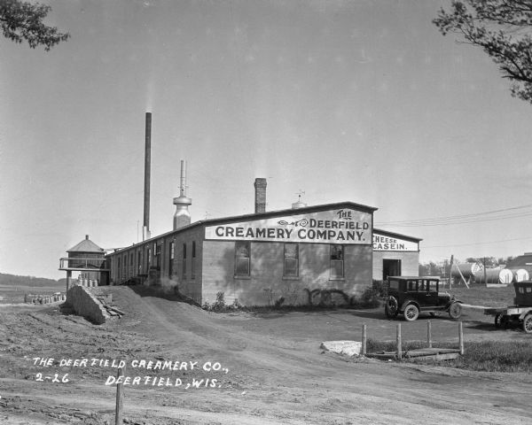 Exterior view of the Deerfield Creamery Company. Two cars are parked in the lot in front of the factory. There are storage tanks in the background on the right, and there appears to be a water tank or tower on the left side of the factory.