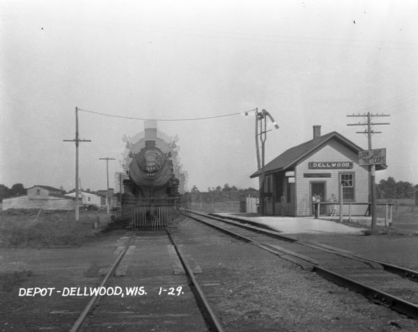 View down railroads tracks of train arriving at the Dellwood Depot. The movement of the train has caused multiple ghost images of the locomotive. A small group of people waits in front of the depot.