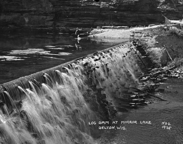 Elevated view over dam and waterfall of a man in a bathing suit walking in the placid and shallow water above the dam. Behind him a boat is tethered along the opposite shoreline of the lake.