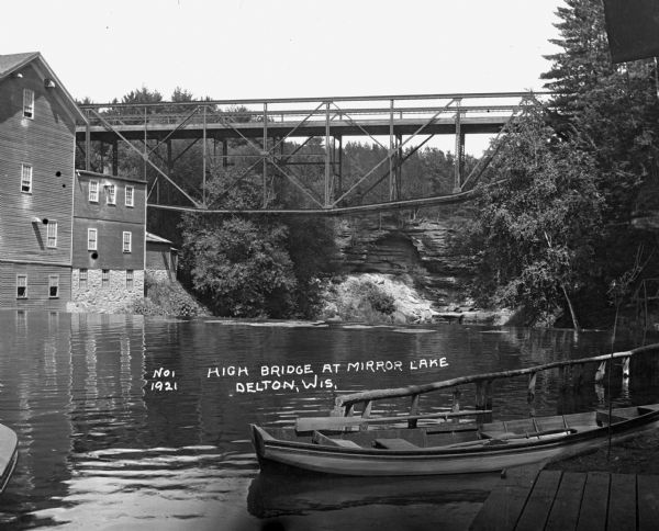View across Mirror Lake. A deck truss bridge crosses the lake high over the dam. Timme Mill, on the left side of the lake, is connected to the bridge. In the foreground, a boat is tied to a wooden dock. In the background, steep cliffs are along the shoreline and covered by trees and plants.