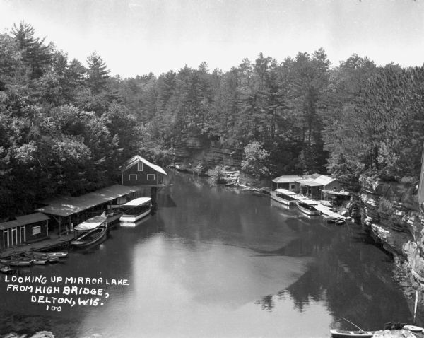 An elevated view of Mirror Lake from a deck truss bridge. On both sides of the lake are boathouses and tour boats. Steep cliffs lined with trees and plants form the shoreline.