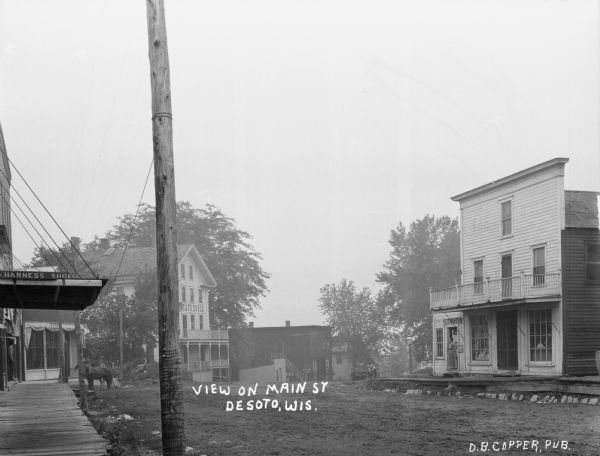 Main Street scene. The shops include a harness shop, bank, drug store, and the Bay State House. A woman stands in the open doorway of the drugstore across the streete, and young boys stand on the sidewalk further down the street.
