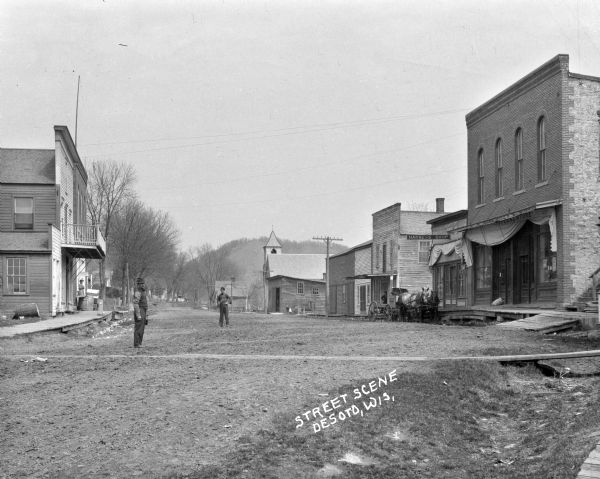 View down street, with two men posed standing in the street. There are wooden sidewalks on both sides of the street, with a storefront on the left, and a woman standing underneath a sign that says: "Butterick Patterns". On the right is a harness shop and a general store. In the background can be seen the Methodist Episcopal Church, and a steep hill rises above it in the far background.