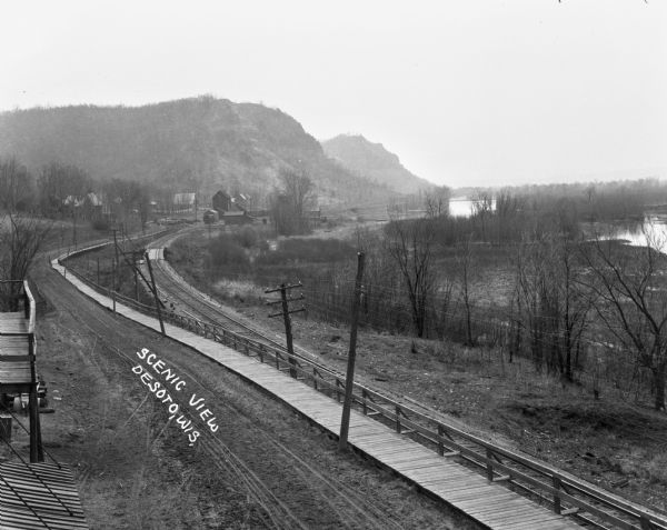 Elevated view of a road, wooden boardwalk and railroad tracks leading towards a group of buildings near bluffs in the distance. On the right is a marshy area along a river, and in the left foreground is the roof and balcony of a building.