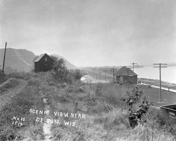 View down path along rough road towards a building labeled "No. 4". The De Soto Depot, railroad tracks, a boxcar, and the Mississippi River are below on the right. In the far background are bluffs.