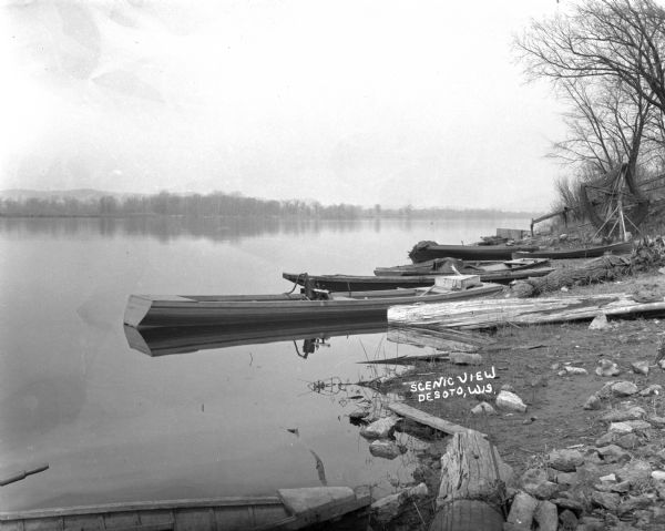 View of shoreline of boats tied along the banks the Mississippi River. A fish net drying reel is near the boats and a wooden walkway. The far shoreline is tree-lined, and a long ridge is in the far background.