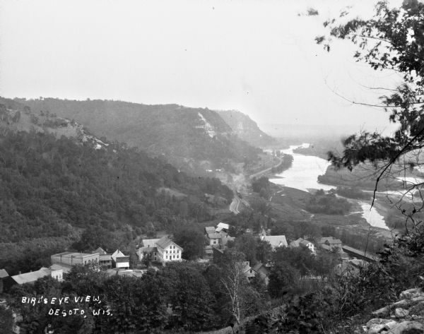 Bird's-eye view from bluff. Far below is Main Street and the Mississippi River. The Bay State House facade can be seen among the buildings lining Main Street. Along the river is a road, and a train is coming along the railroad tracks. Dwellings, church buildings and fields can be seen among the trees.