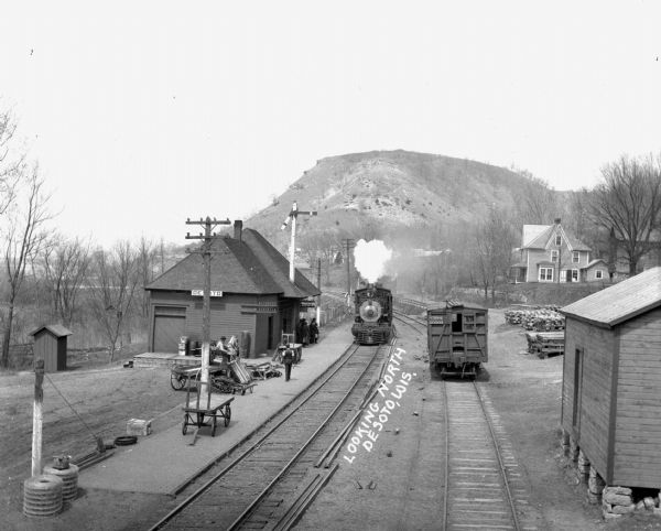 Elevated view of the De Soto depot. A locomotive, blowing steam, is approaching the platform, where a small group of people waits to board. A boxcar on the side track has the identifying marker, "CB&Q 60003." The Mississippi River is on the left, and a steep bluff is in the background.