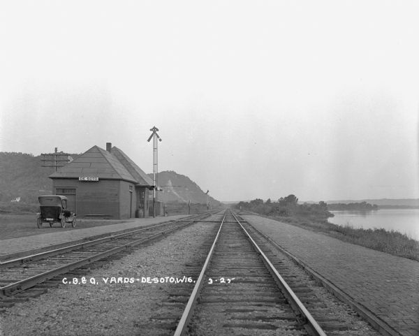 View down double set of railroad tracks with the De Soto depot on the left and the Mississippi River on the right. A car is parked at the depot, and bluffs are in the background. There are brick walkways lining both sides of the tracks.