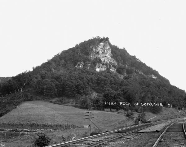 View across double set of railroad tracks and field towards steep bluff with exposed rock.