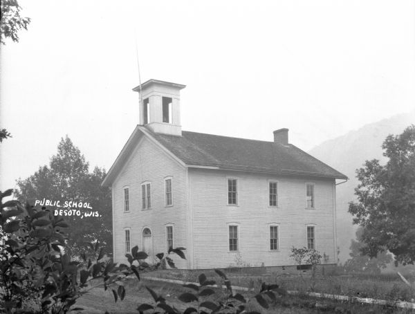 Exterior of the two-story public schoolhouse with a bell cupola and flag pole. There is a wooden sidewalk in front of the school building. Branches and leaves in the foreground obscure the view of the school. There is a bluff in the background on the right.