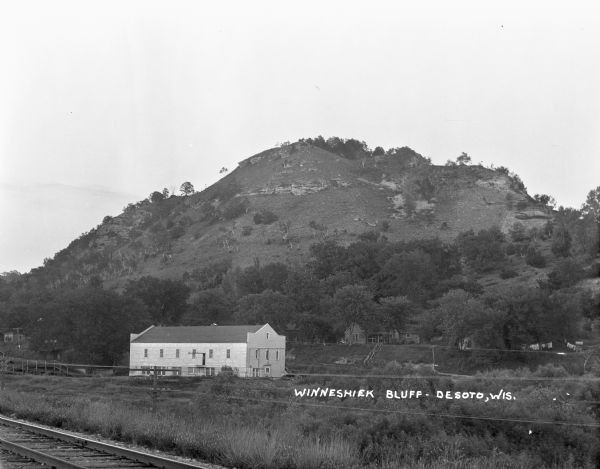 View near railroad tracks of a two-story industrial building and houses at the base of a bluff.