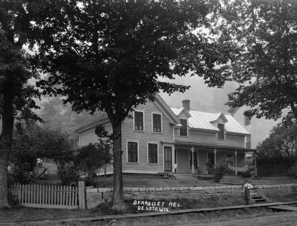 Exterior of the Beardsley's house from the street. A young man in overalls stands on the wooden sidewalk. There is a hammock on the porch. A large building of stone or brick is on the right, and in the background there appears to be a tree-lined bluff.