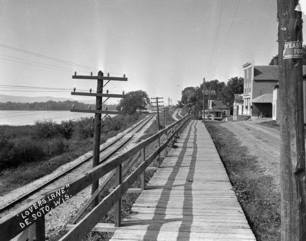 View down a wooden pedestrian walkway. The Mississippi River and railroad tracks are along the shoreline on the left. Storefronts and other buildings are along a road on the right. An advertisement for Yeast Foam is posted on the utility pole in the foreground.