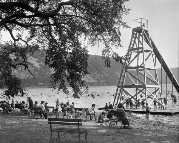 View looking out at beach scene at Devil's Lake. There are benches on the shore where vacationers observe the swimmers. Two wooden structures have ladders up to slides in the water. A large slide is on the shoreline, and a smaller slide is out in the lake.