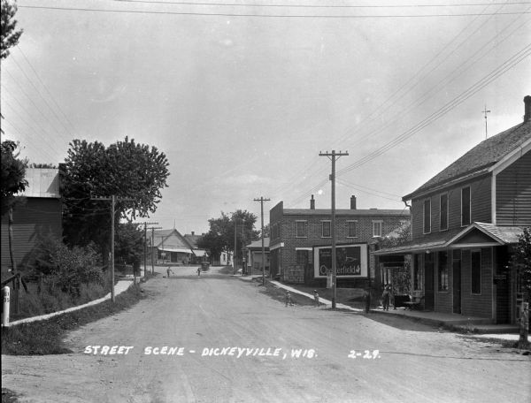 View down street with children playing in the background. Along the street is a row of businesses and storefronts. Advertisements for Chesterfield cigarettes and Meuser Lumber Company are on the right. D.W. Lawrence General Merchandise store is at the intersection in the background.