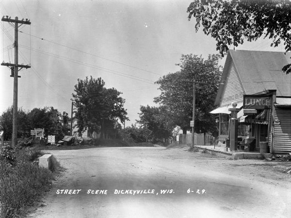 View down unpaved street towards the D.W. Lawrence General Merchandise store, which has a gas pump in front of the storefront. Two automobiles are parked on the left in the grass near numerous roadside signs.