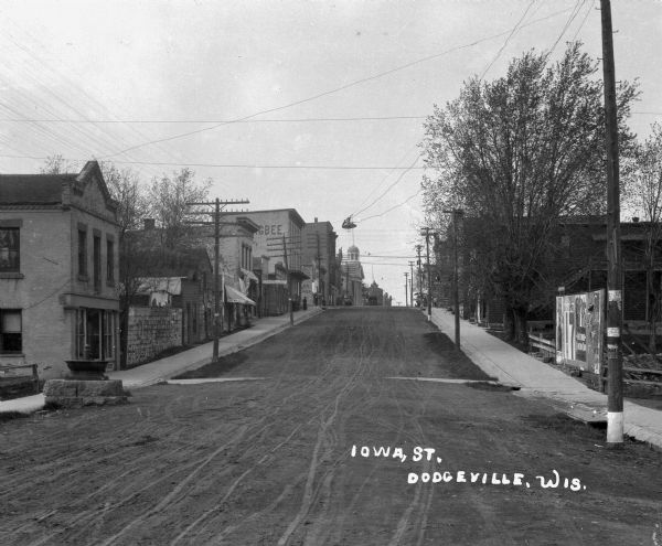 View up unpaved street. There is a horse-drinking fountain on the left side of the road, and pedestrians are on sidewalks in the background.