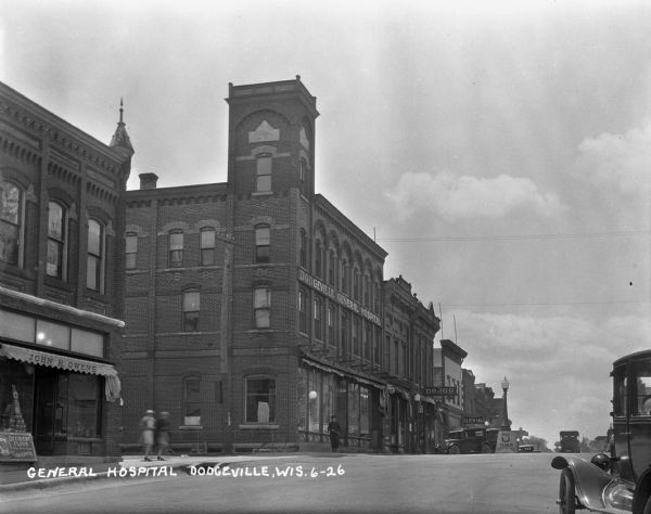 View across street of the Dodgeville General Hospital on the corner of Chapel and Iowa Streets. Along Iowa street, there is a John H. Owen's general store, a drugstore, and a clothing store. There are parked automobiles, and pedestrians are walking along the sidewalk.