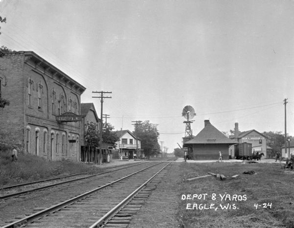 View down railroad tracks towards the Eagle train depot. A number of pedestrians are around the depot. On the left near the road, two men stand in front of a restaurant. A woman stands in front of another  building under a sign that says: "Ice Cream Parlor."